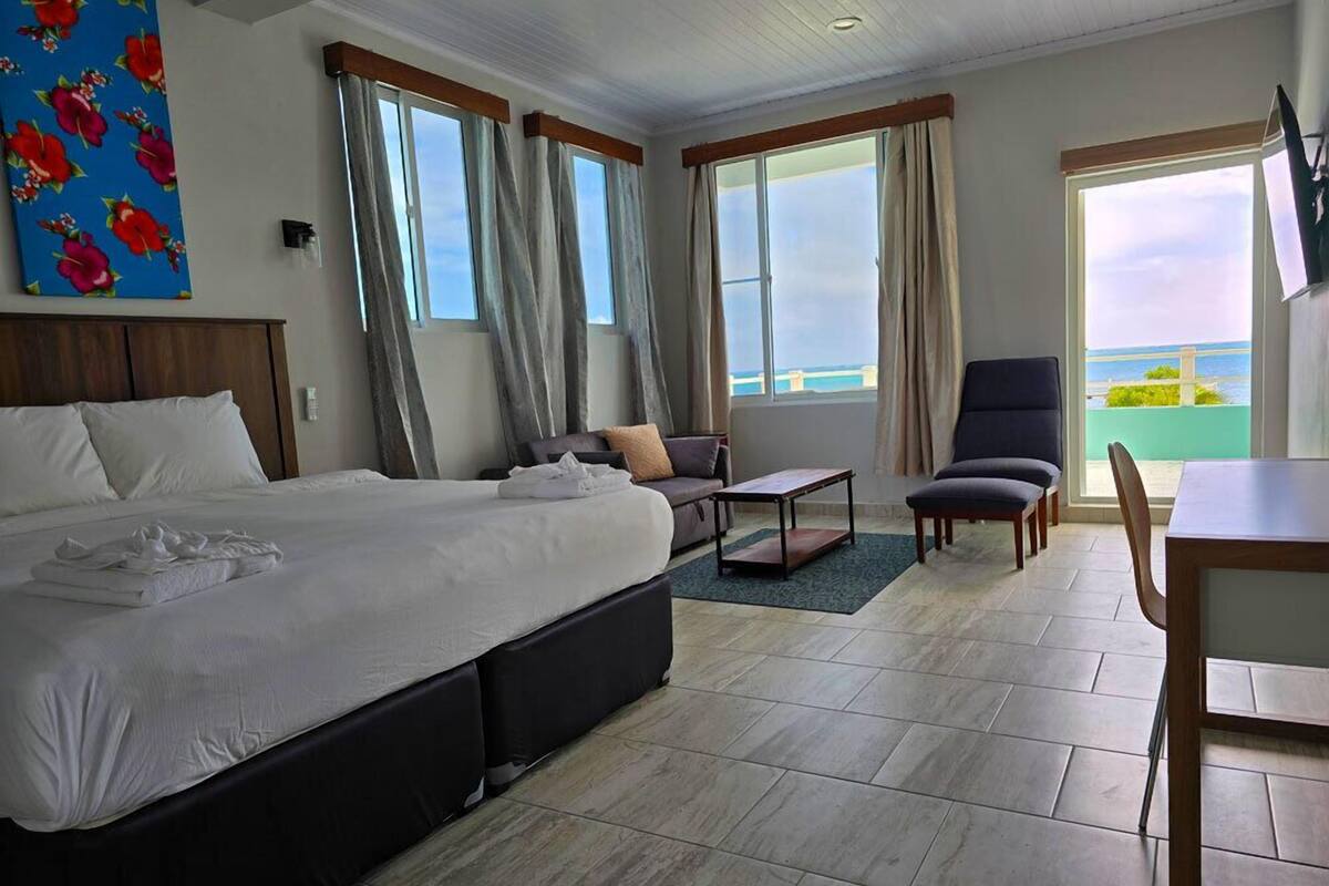 Ein Hotelzimmer mit einem großen Bett, einer Couch, einem Esstisch und einem Stuhl. Es gibt Fenster mit Vorhängen, ein Gemälde an der Wand und Meerblick.