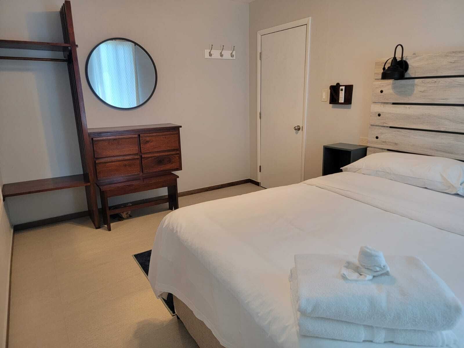 A hotel room with a bed, a wooden dresser, a mirror, a door, and a bedside table with a phone.