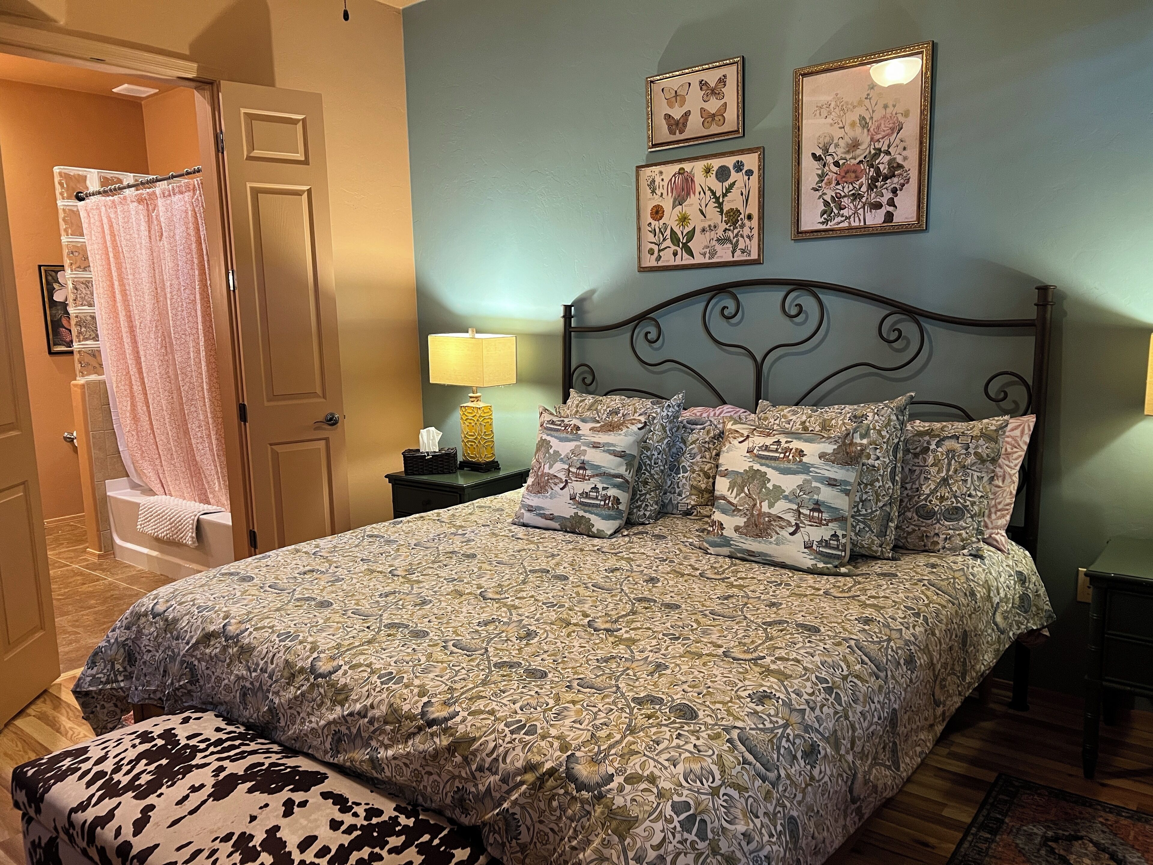 Ein Schlafzimmer mit einem gemusterten Bettbezug, einem Kopfteil und dekorativen Kissen. Ein Nachttisch mit Lampe und ein botanisches Bild in einem Rahmen hängen an der Wand.