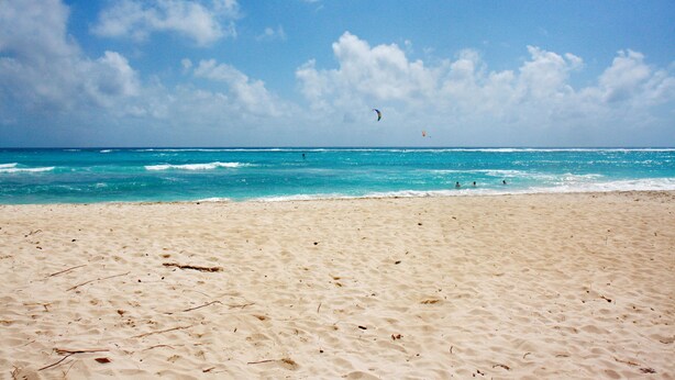 Uma praia arenosa com águas azul-claras e algumas pessoas ao longe.