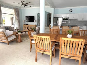 A spacious living area with a wooden dining table, chairs, and a sofa. A TV is mounted on the wall, and there is a kitchen area with cabinets and appliances.