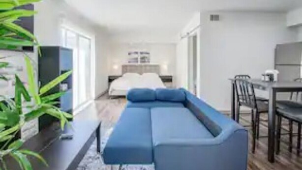 A modern hotel room with a large blue sectional sofa, a dining area with a table and chairs, and a bed in the background.