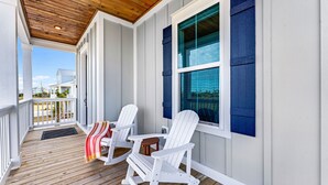 A covered porch with two rocking chairs and a small table, wooden deck flooring, and a view of the ocean and other buildings in the background.
