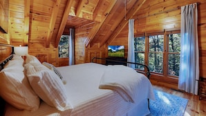 Un dormitorio de cabaña de madera con una cama grande, un televisor y vistas al exterior.