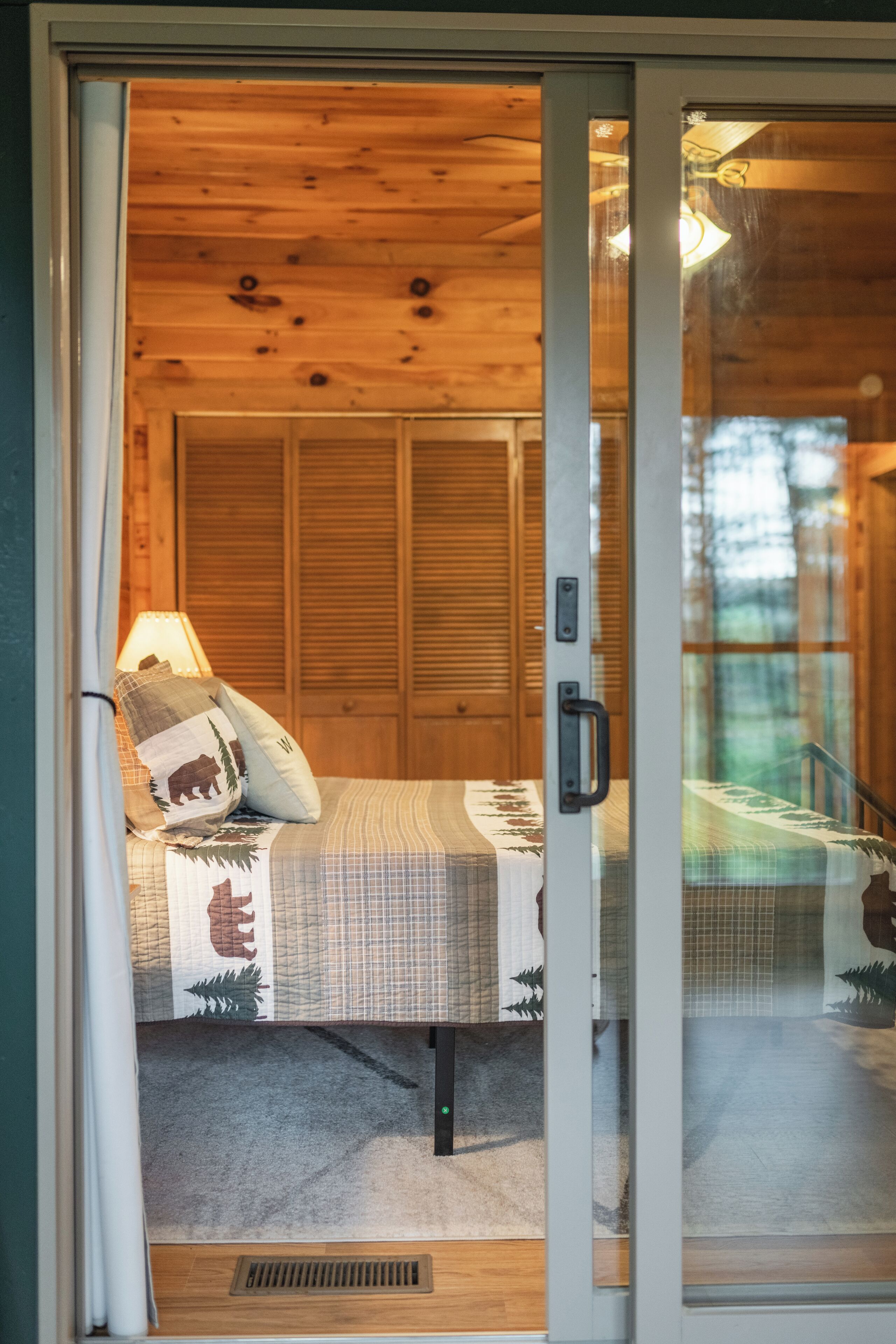 A wooden cabin interior with a bed, a lamp, and a view of the outside.