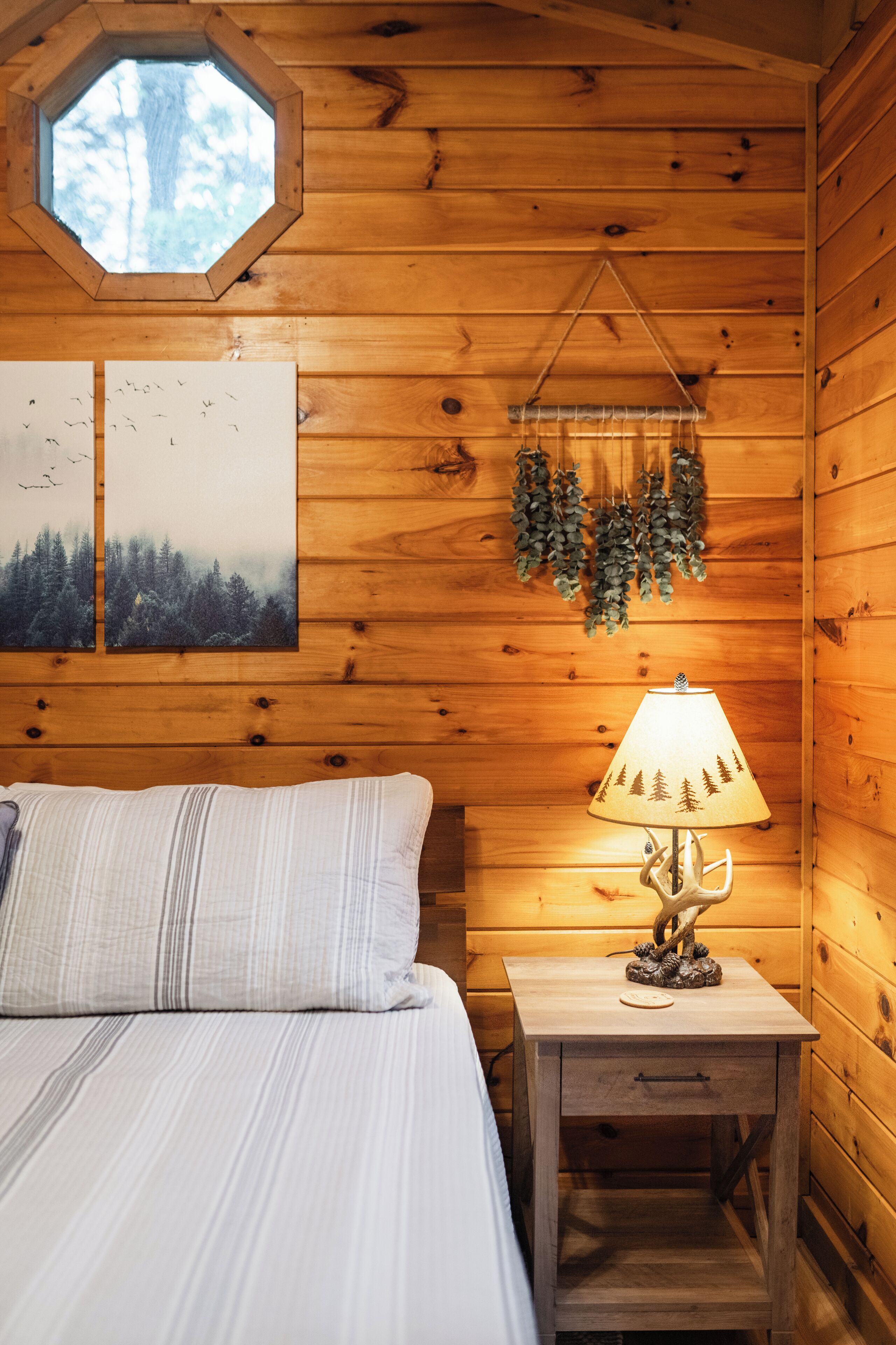 A wooden cabin room with a bed, a nightstand with a lamp, a window, and wall art.
