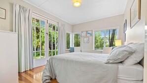 A bedroom with a large bed, a nightstand, a lamp, and a view of greenery through the windows. - Fisherman’s retreat in Turangi (Turangi)
