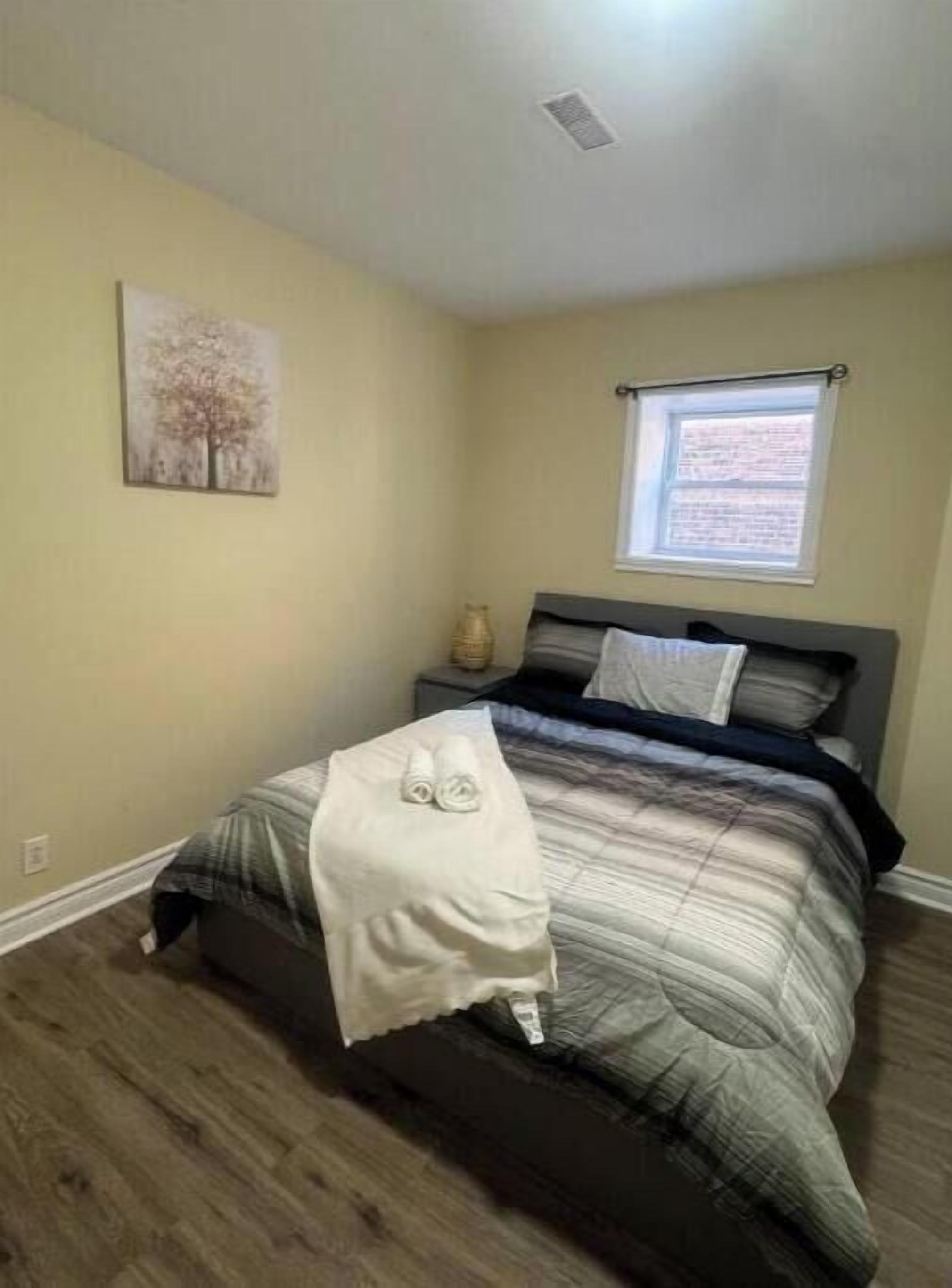 Un dormitorio con cama, mesita de noche, ventana y un cuadro en la pared.