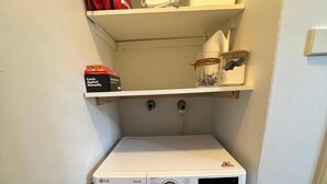 Een kleine wasruimte met een voorlader wasmachine, een plank erboven met een witte bak, een rode tas en een doos met de tekst 