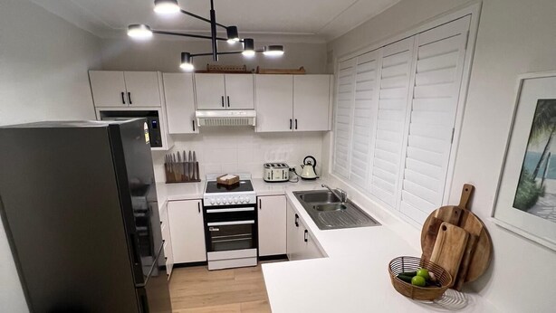 Een compacte keuken met witte kastjes, een inbouwoven, een spoelbak en een klein eethoekje met twee stoelen.