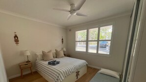 Een slaapkamer met een bed, nachtkastje, raam met zonwering en plafondventilator.