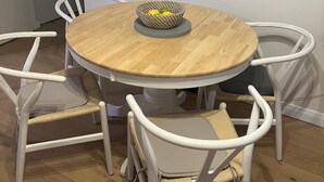 Een ronde houten eettafel met een gevlochten schaal erop, omringd door vier witte stoelen met rieten zittingen.