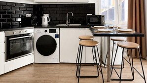 Een moderne keuken met witte kasten, een zwart betegelde spatwand, een wasmachine, een magnetron en een eethoek met krukken.