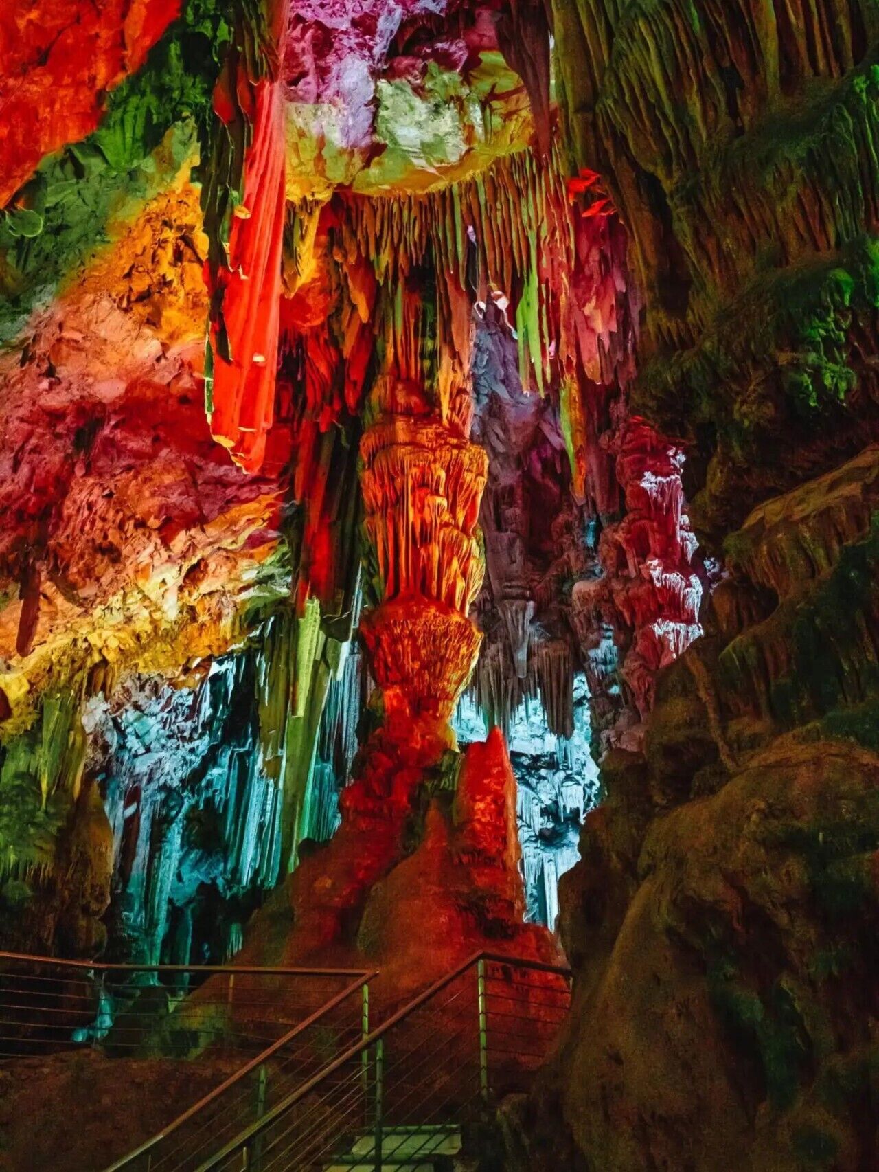 Una cova amb estalactites i estalagmites de colors, una barana de metall i una escala que baixa.