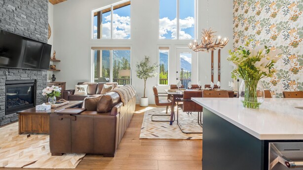 A spacious living room with a fireplace, large windows, a dining area, and a kitchen with a central island. - Spring Creek 3BR Penthouse ~ Wide Mountain Views ~ (Canmore)