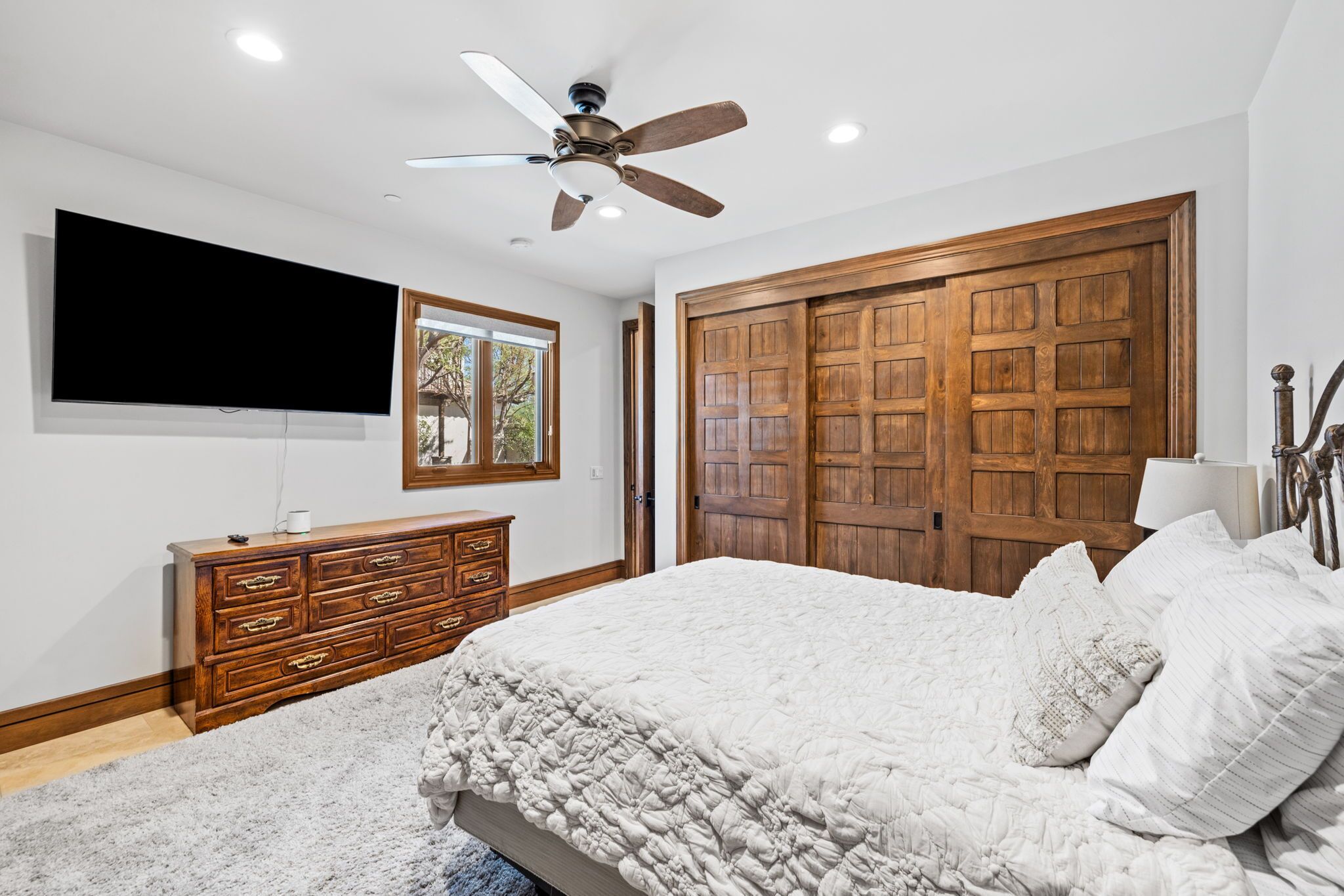 A bedroom with a large bed, wooden wardrobe, ceiling fan, TV, and a window with a view.