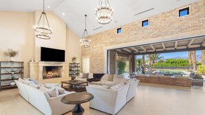 A spacious living area with a fireplace, sofas, a TV, and a large window overlooking a garden. - 3.5 Acre compound- 2 pools, tennis, pickleball, basketball, sauna & steam + more (Bermuda Dunes)