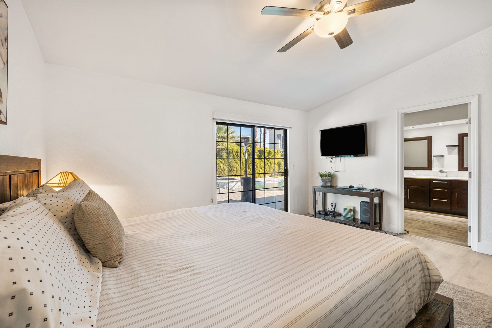 A bedroom with a bed, a ceiling fan, a TV, a window, and a bathroom visible through an open door.