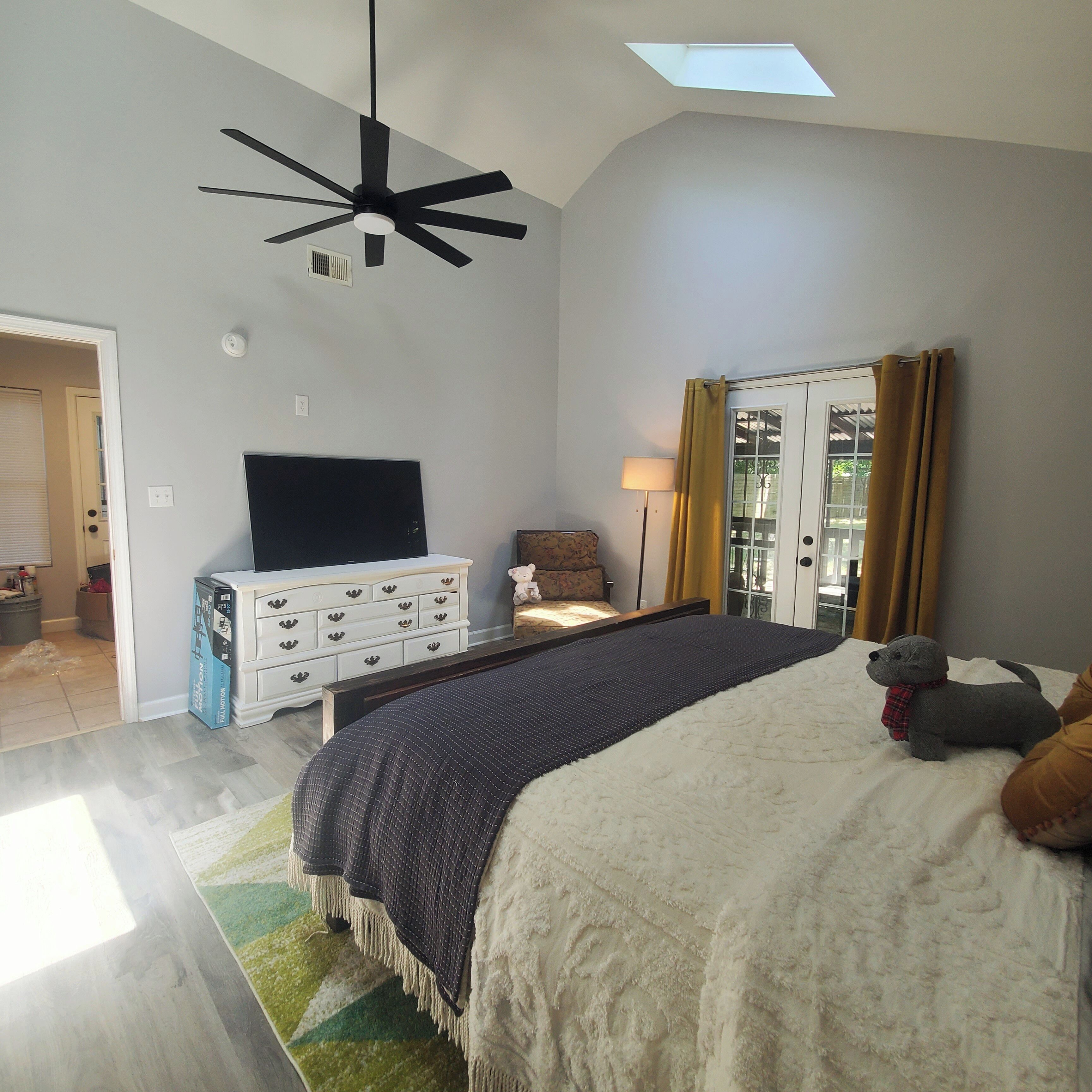 Une chambre avec un grand lit, un ventilateur de plafond, une télévision, une commode et une fenêtre avec des rideaux.