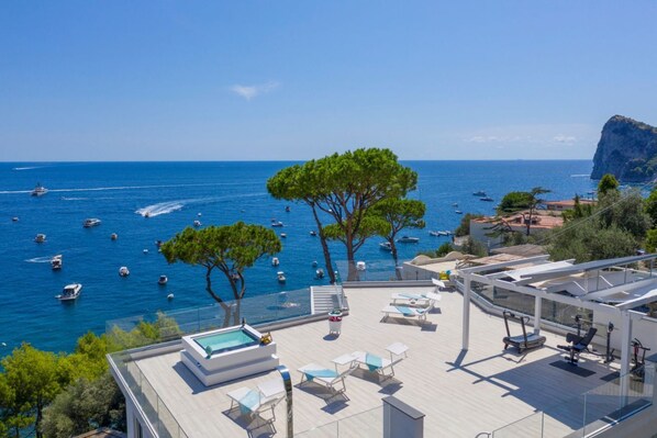 Un área de piscina en la azotea con sillones de descanso, un jacuzzi y vistas al mar con barcos.
