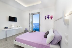 A modern hotel room with a bed, a desk, a TV, and a window with a view of the sea.