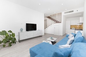 A modern living room with a flat-screen TV, a blue sofa, a coffee table, and a staircase leading to a kitchen.