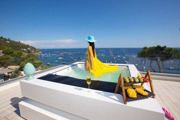 A woman in a yellow dress and blue hat sitting in a hot tub with a view of the sea and boats.