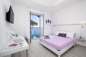 A bedroom with a bed, a desk, a TV, and a view of the sea.