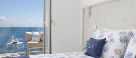 A bedroom with a bed, pillows, a nightstand, a chair, and a view of the sea.