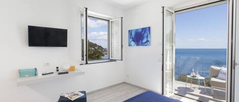 A modern bedroom with a bed, a TV, a window with a view of the sea, and a balcony with a table and chairs.