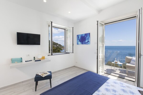 A modern bedroom with a bed, a TV, a window with a view of the sea, and a balcony with a table and chairs.
