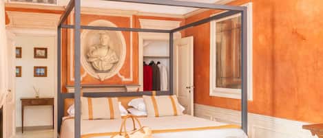 A four-poster bed with a beige and white color scheme, a small table with a cup and saucer, and a bust sculpture on the wall.