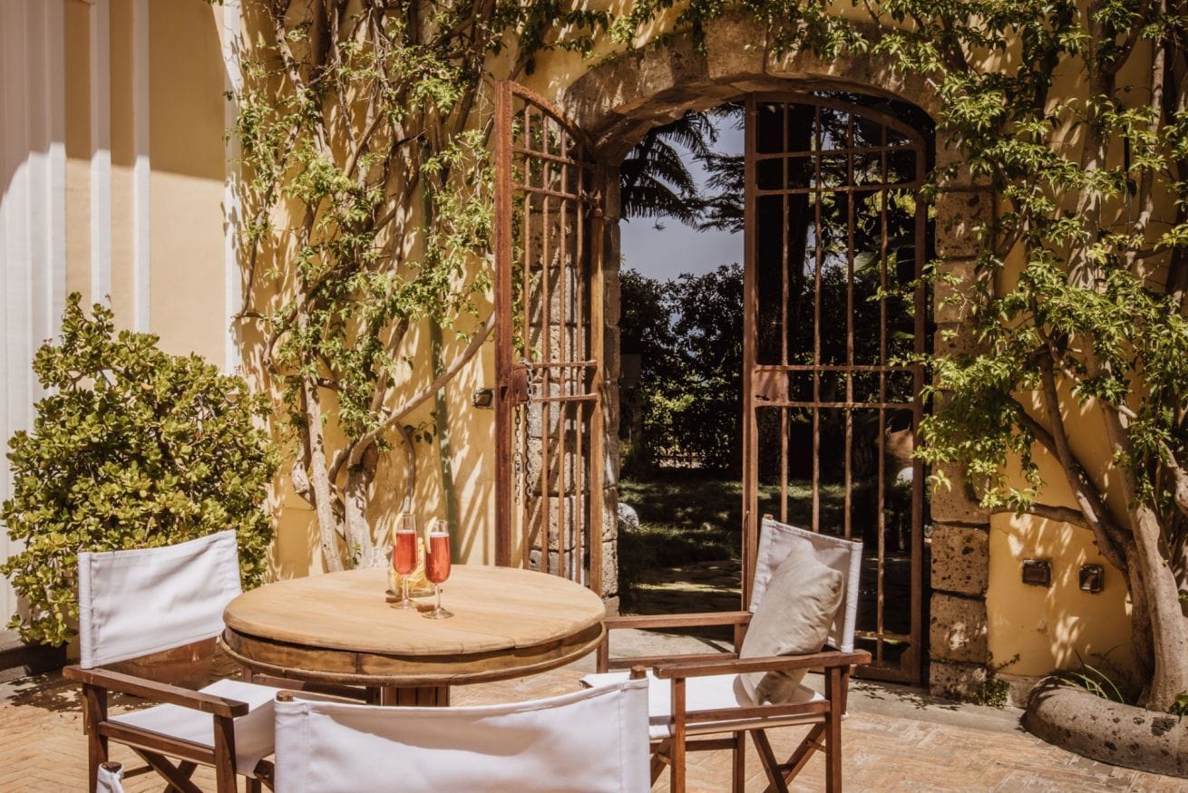 An outdoor patio with a round wooden table and four chairs, surrounded by lush greenery and a stone wall.