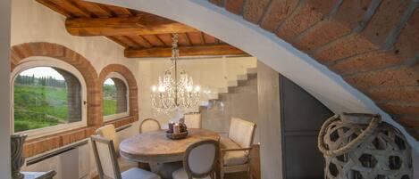 A dining area with a round table, chairs, and a chandelier. There are arched windows and a brick wall.