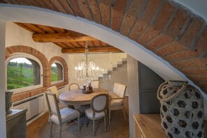 A dining area with a round table, chairs, and a chandelier. There are arched windows and a brick wall. - MyExclusiveItaly - Chianti's Villa | Vineyard, infinity pool, view (Greve in Chianti)