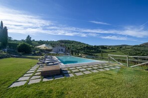 A pool area with lounge chairs, a wooden deck, and a view of a hill with a house in the background. - MyExclusiveItaly - Chianti's Villa | Vineyard, infinity pool, view (Greve in Chianti)