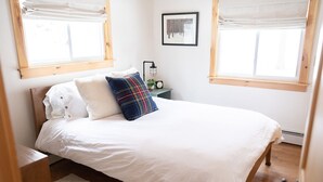 Ein Schlafzimmer mit einem Bett, zwei Fenstern mit Jalousien, einem Nachttisch mit Lampe und einem gerahmten Bild an der Wand.