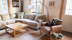 Ein gemütliches Wohnzimmer mit einem Ecksofa, einem kleinen Holztisch, einem Ledersessel und einer Fußkorb mit Hausschuhen.