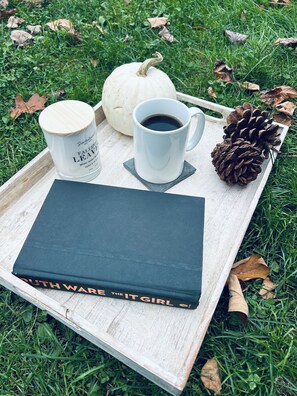 Una bandeja con una taza blanca, una vela blanca, un libro titulado 'GOTHAM IT GIRL', una calabaza blanca y una piña sobre una superficie de madera con césped y hojas caídas alrededor.