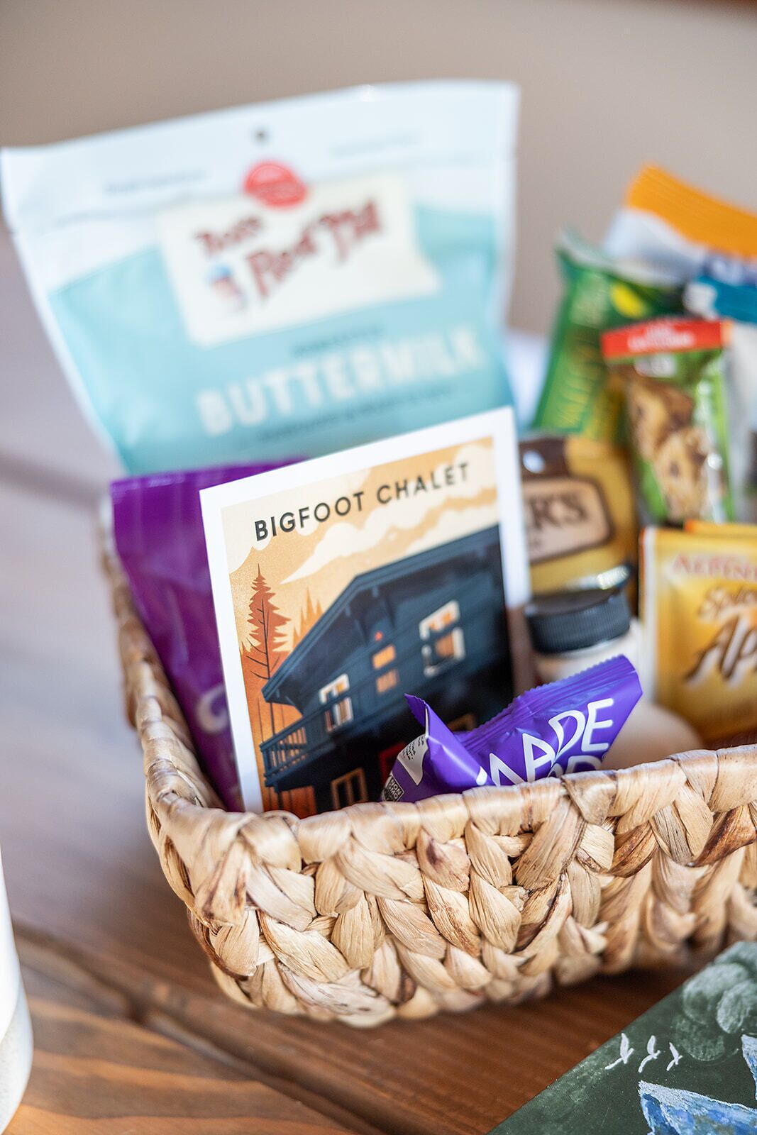 A basket containing a variety of food items including a bag of Buttermilk, a package of Bigfoot Chalet, and other snacks.