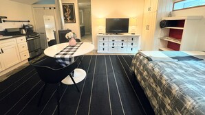 A compact hotel room with a kitchenette, a dining table with a checkered table runner, a flat-screen TV, a bed with a patterned duvet, and a small bookshelf. - Private Cozy Garden Studio Retreat - Near Downtown, Gardens & Forest Park (Portland)