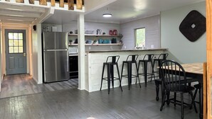 A modern kitchen with a stainless steel refrigerator, a microwave, and a sink. There are bar stools and a wooden dining table. - The ‘A’mazing Escape! Beaches, hiking trails, & great fall color tour location! (Manistee)