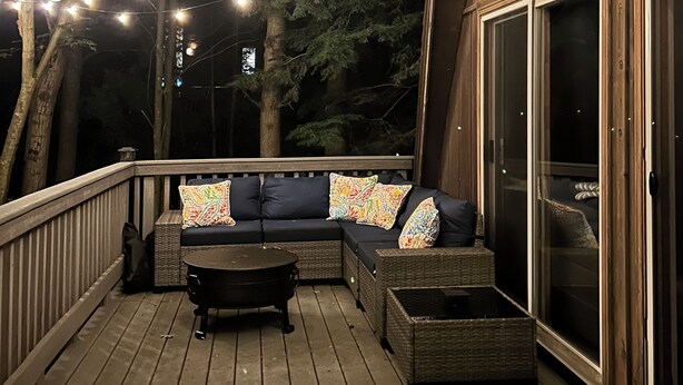 A wooden deck with a dark-colored sofa and a small table, surrounded by string lights. - The ‘A’mazing Escape! Beaches, hiking trails, & great fall color tour location! (Manistee)