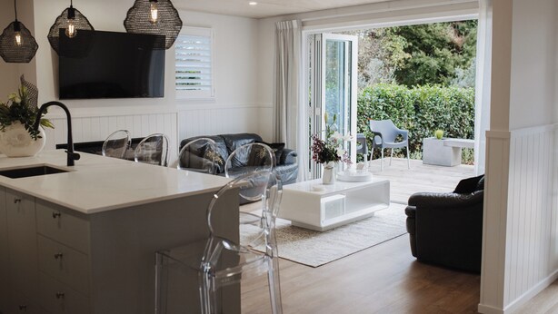 A modern kitchen with a central island, a dining area with chairs, a sofa, and a TV mounted on the wall. - The Doctors Villa Waikanae (Waikanae Beach)