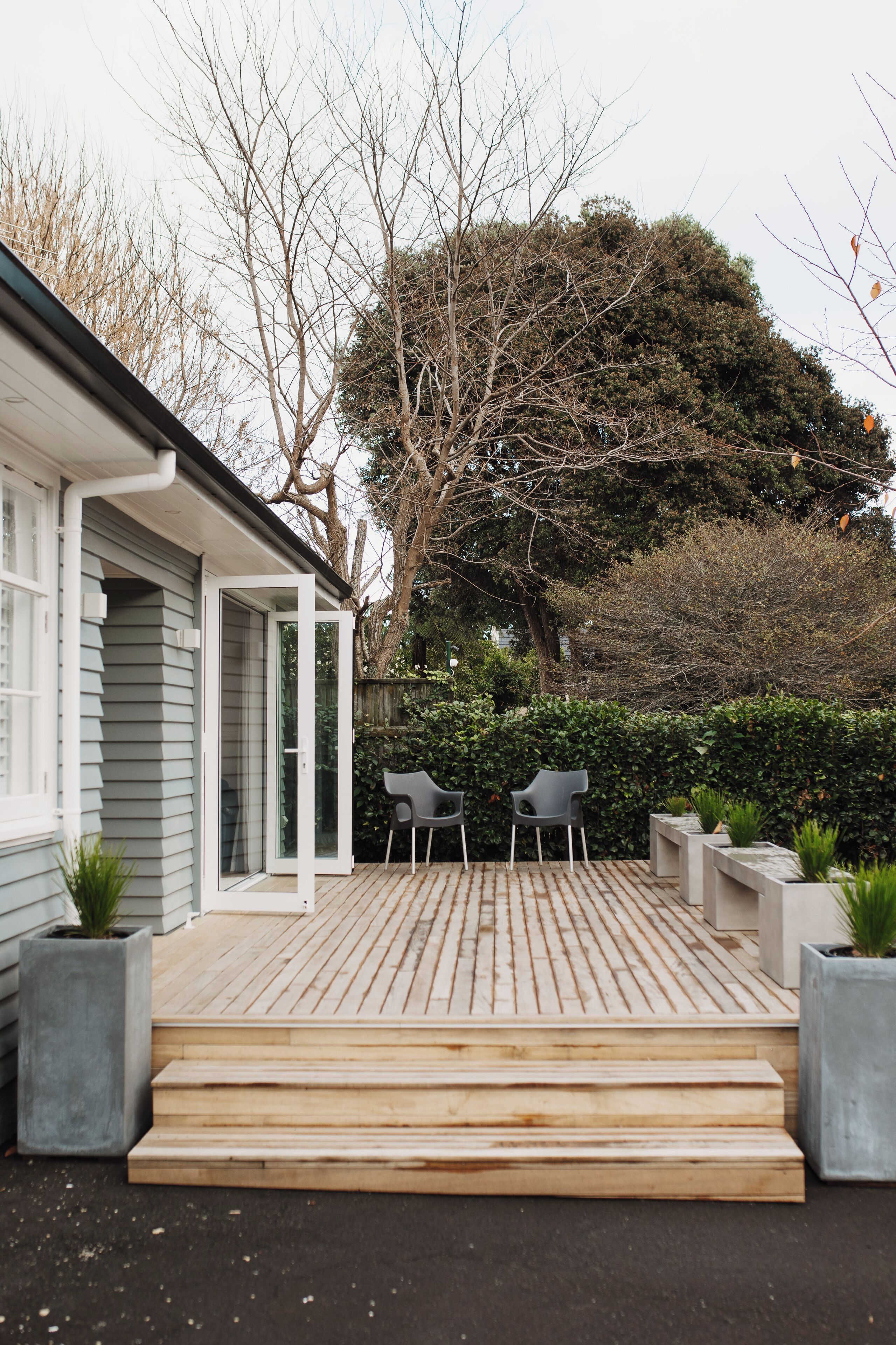 A modern house with a wooden deck, two chairs, and a large tree in the background.