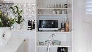 A modern kitchen with white cabinetry, a microwave, coffee maker, and a clear chair. - The Doctors Villa Waikanae (Waikanae Beach)