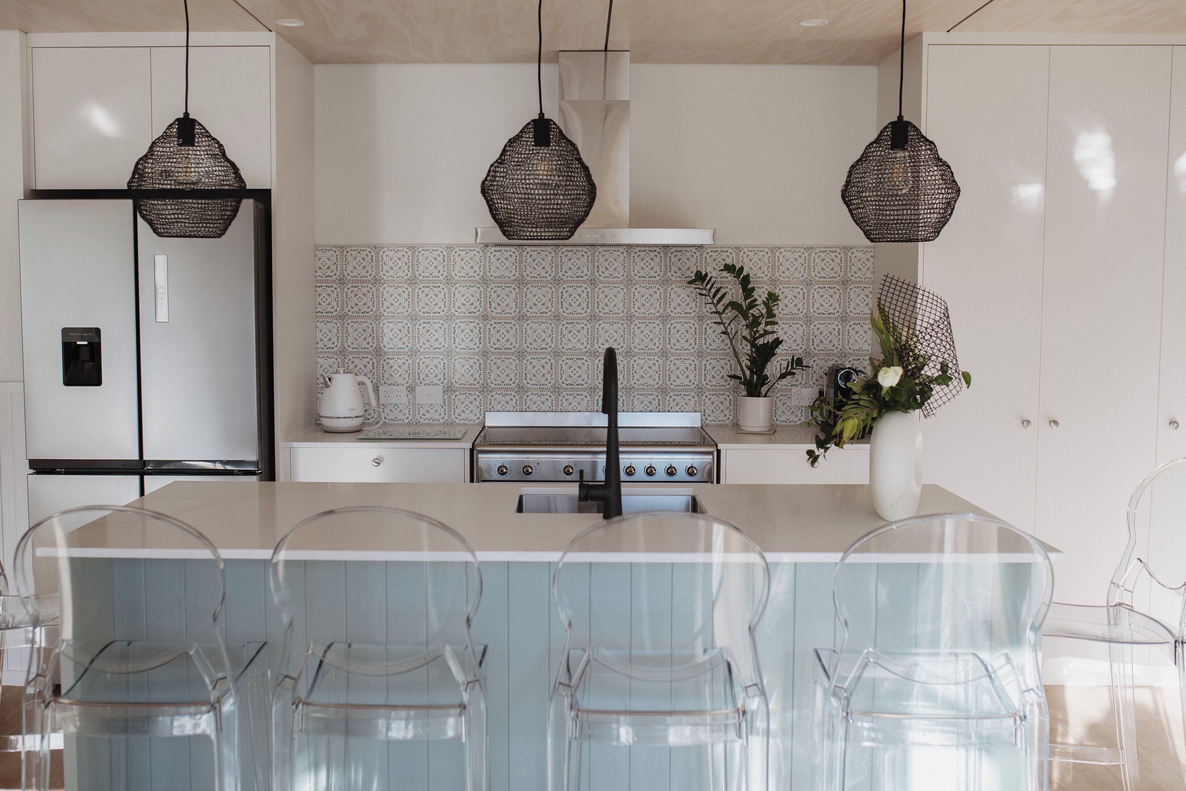 A modern kitchen with a central island, clear chairs, a stove, and decorative hanging lights.