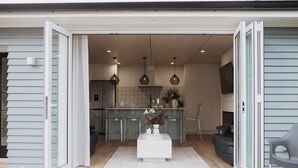An open sliding glass door leading to a modern kitchen with a central island, pendant lighting, and a dining area with a grey sofa. - The Doctors Villa Waikanae (Waikanae Beach)