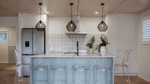 A modern kitchen with a central island, clear bar stools, and pendant lighting. - The Doctors Villa Waikanae (Waikanae Beach)