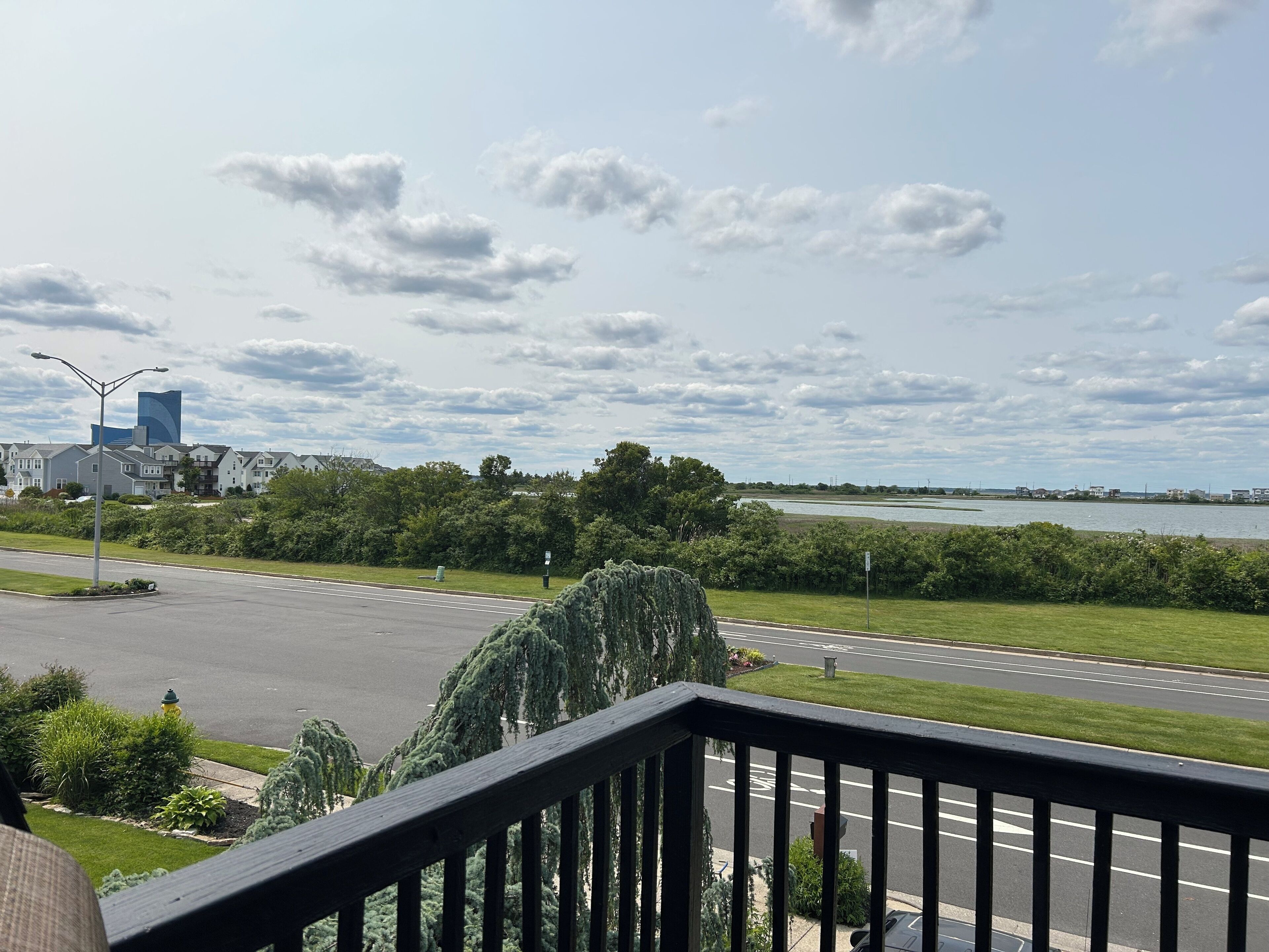 A view from a balcony overlooking a road with a water body in the distance, a parking lot, and a building with a blue structure.